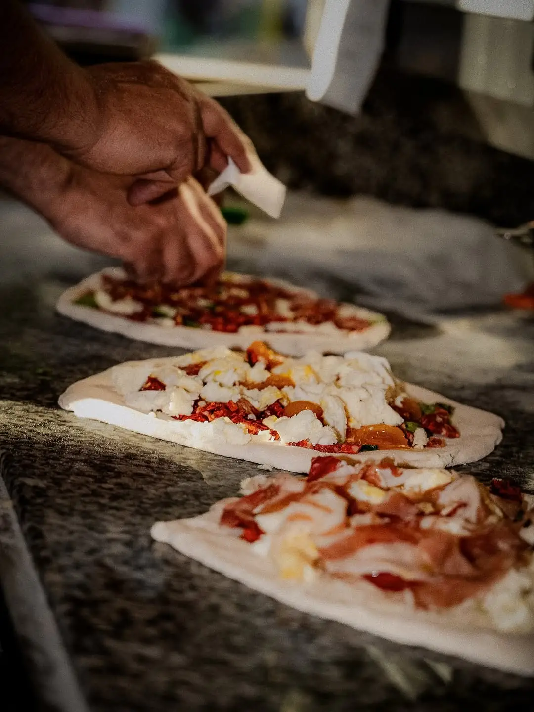 Hände eines Pizzaiolo beim Belegen frischer Pizzen auf der Marmortheke