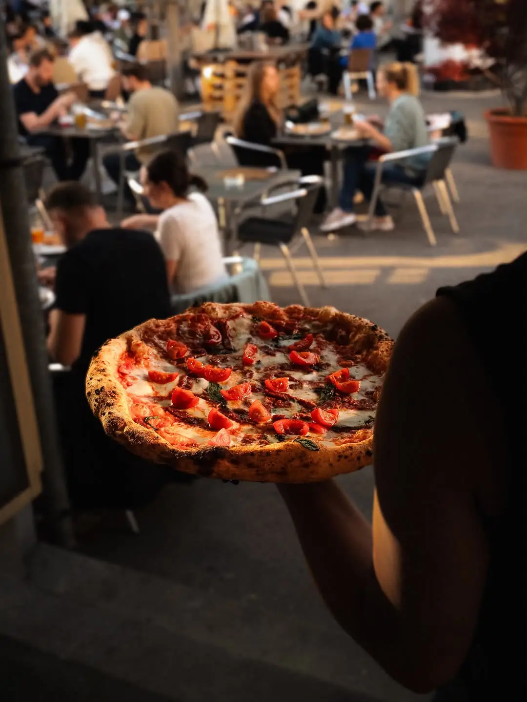Frisch gebackene Margherita wird vor der belebten Gastgarten-Terrasse hochgehalten