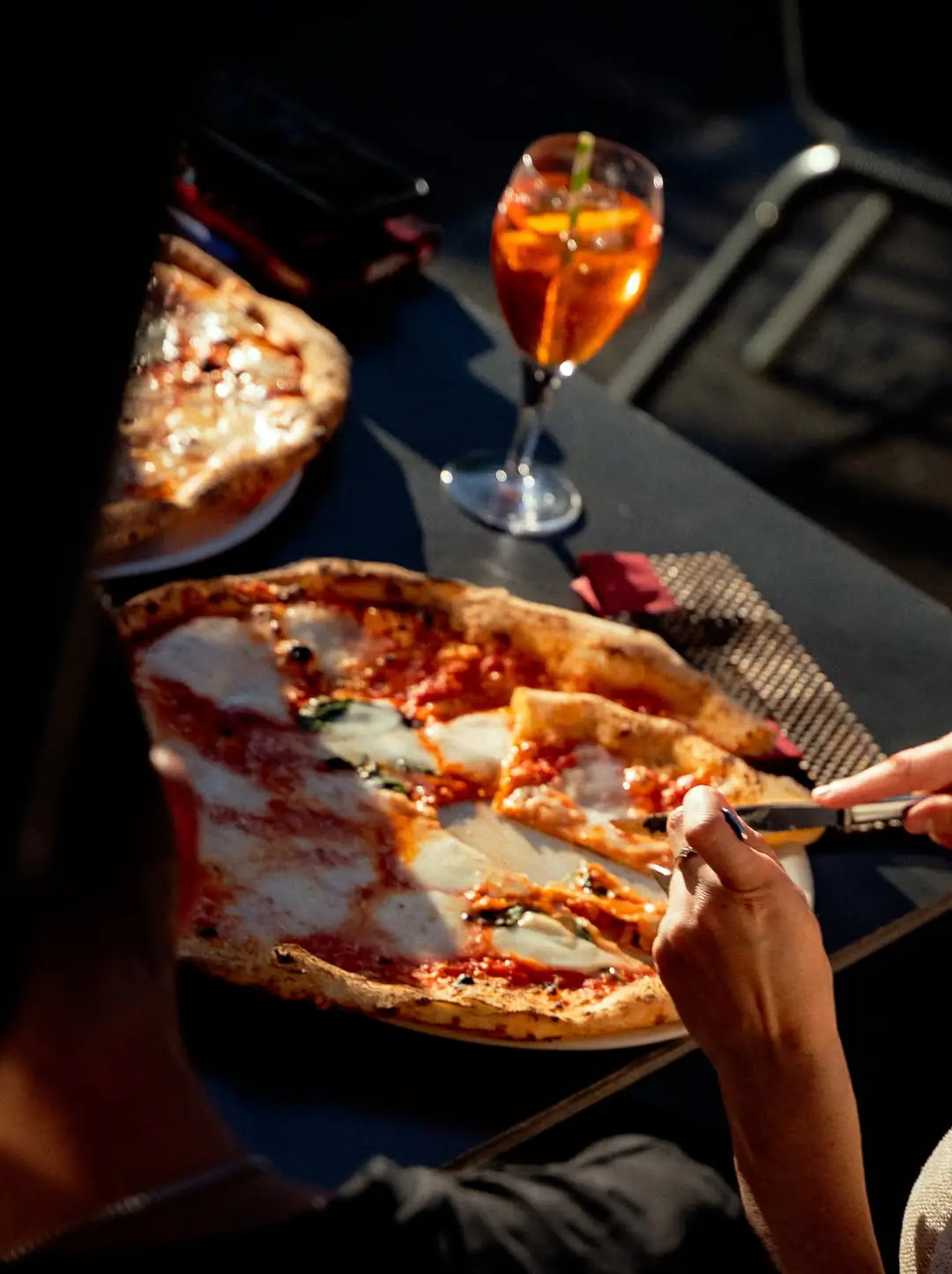 Zwei frisch gebackene Pizzen und ein Spritz auf einem Holztisch draußen in warmem Abendlicht